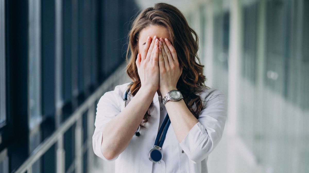 Женщина-медик в голубой форме и перчатках держится за голову; на шее стетоскоп, выражение усталости и стресса