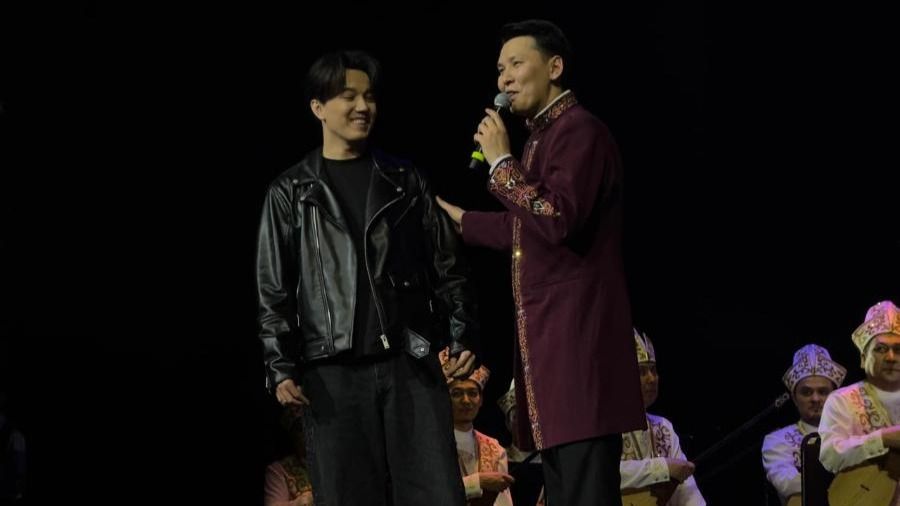 Димаш Кудайберген стоит на сцене рядом с дирижёром Абылаем Тлепбергеном, который говорит в микрофон; на фоне &mdash; музыканты национального оркестра