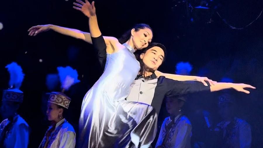 Танцевальная пара исполняет номер на сцене: мужчина поддерживает женщину в светлом платье, на фоне сидят музыканты в национальных костюмах
