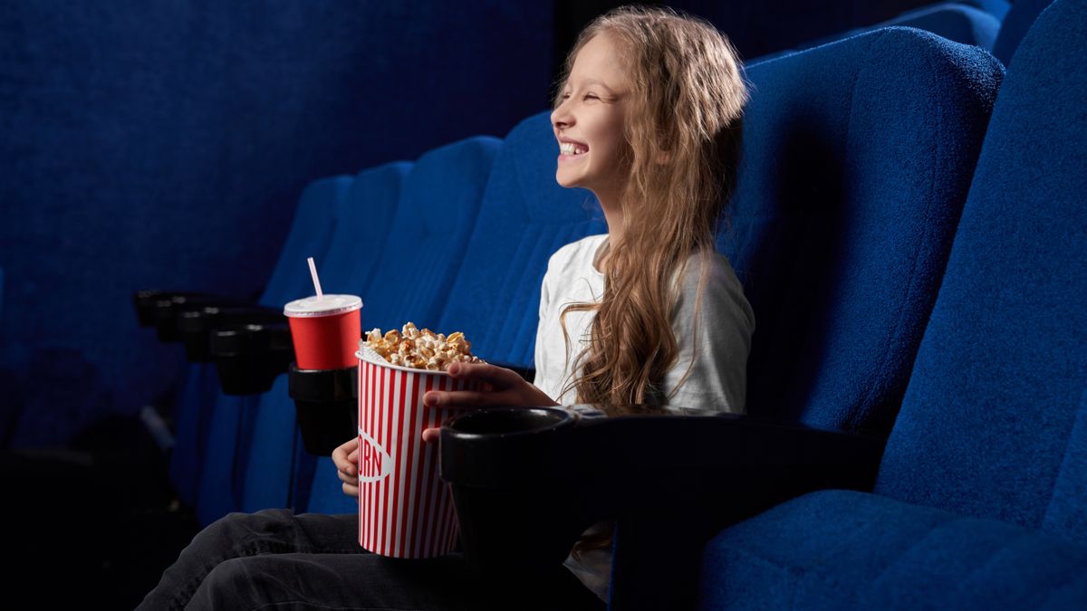 Юная зрительница увлечённо смотрит фильм в кинотеатре с попкорном на руках 
