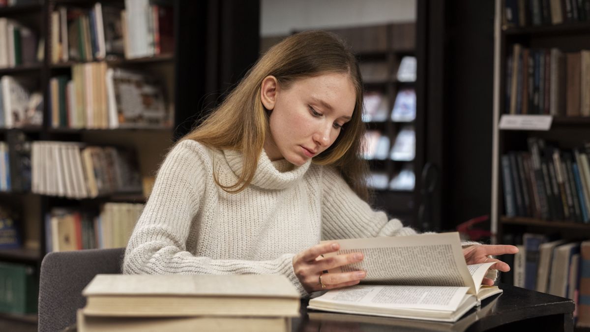 Девушка читает книгу за столом в библиотеке среди книжных полок