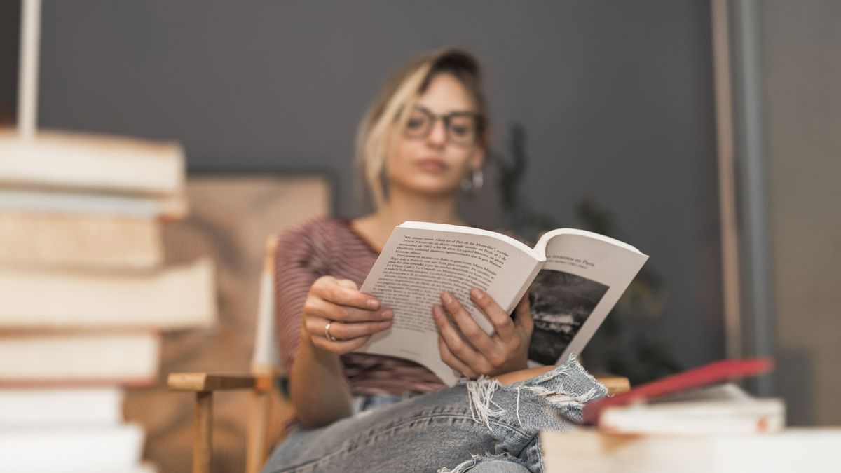 Девушка читает книгу, сидя в кресле среди стопок книг